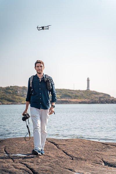 Aykut Argun, photographer and videographer, holding a camera on the rocks by the sea while a drone flies overhead