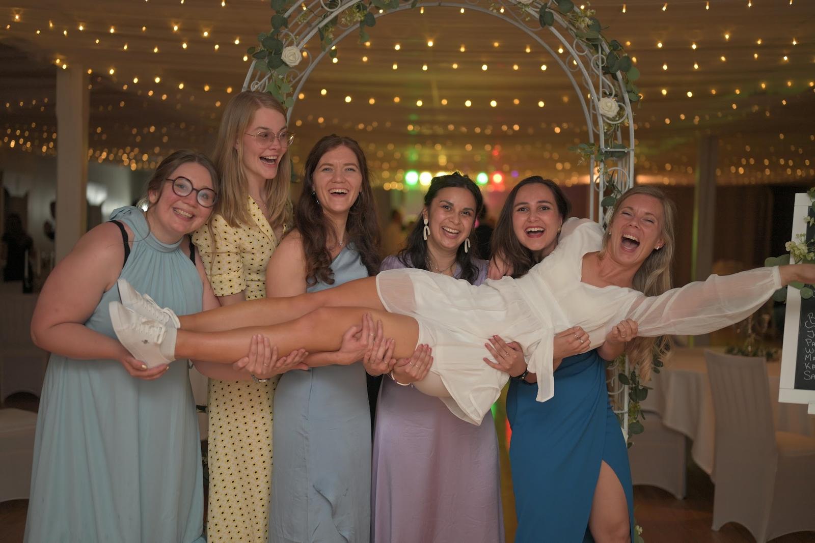 The bride lifted up by laughing friends at the party under an arch of fairy lights