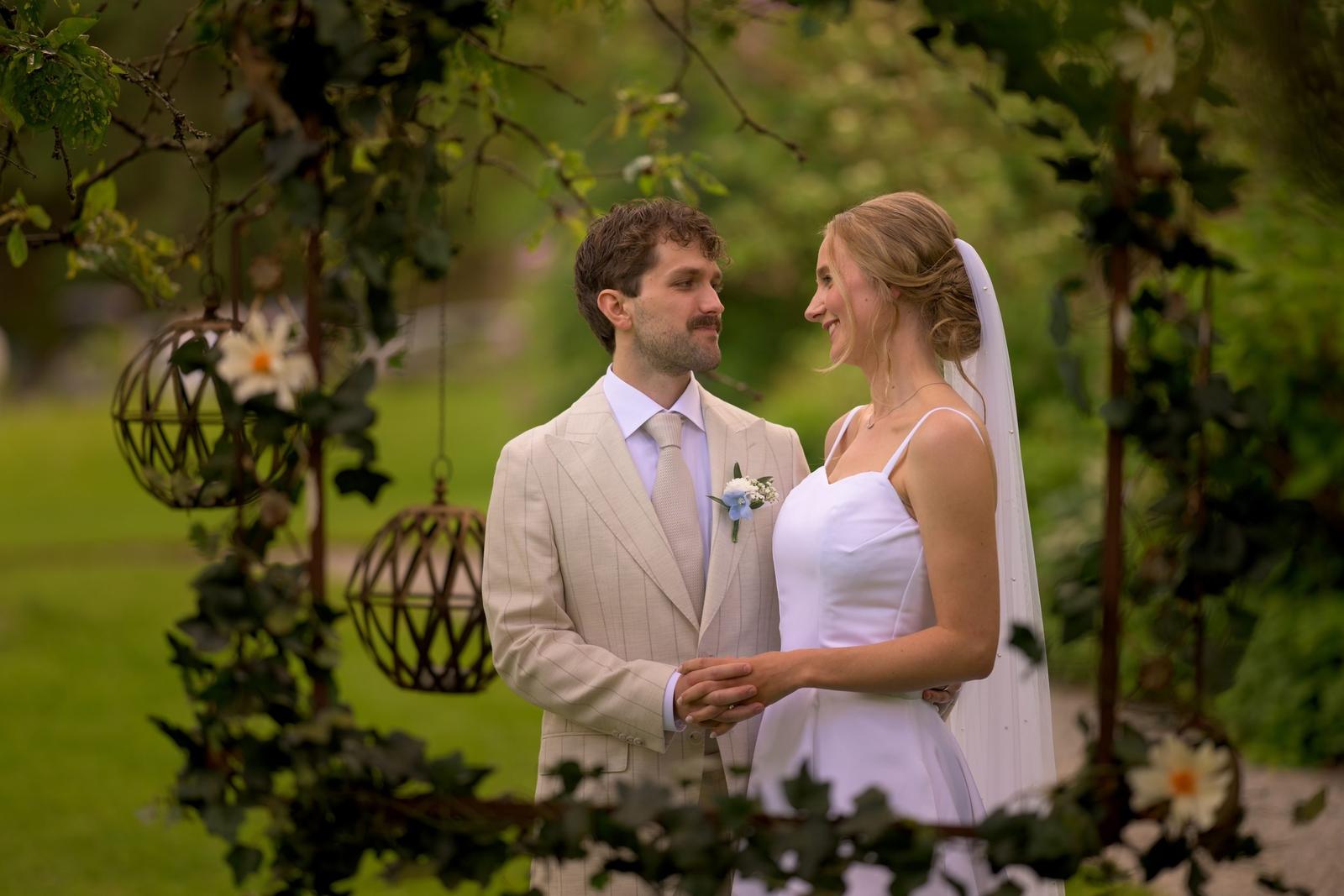 Bride and groom holding hands under an ivy arch with hanging lanterns in the garden at Villa Vanahem in Stenungsund