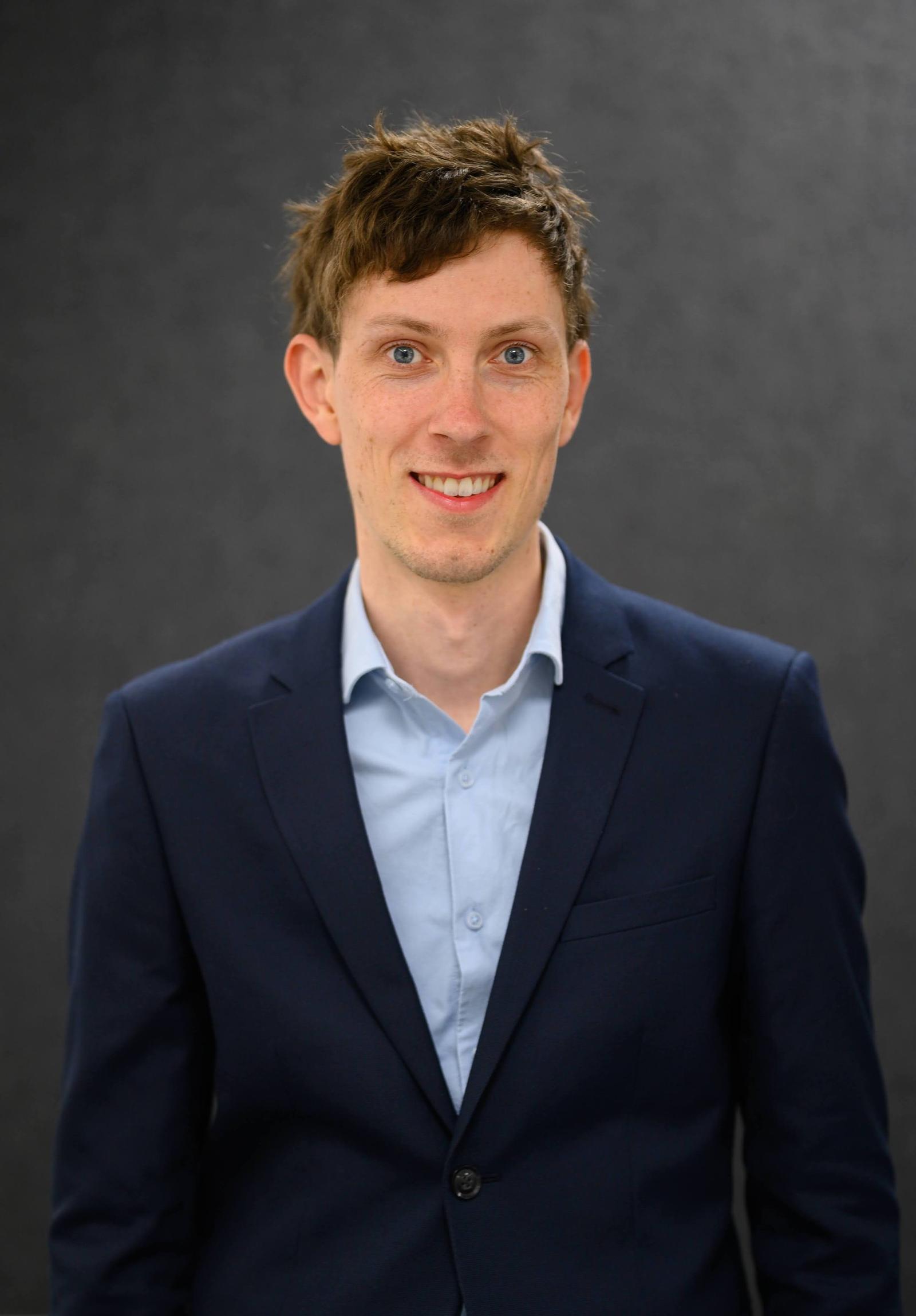 A young man with curly brown hair and blue eyes, wearing a navy blazer and a light blue shirt, smiling in front of a dark background.