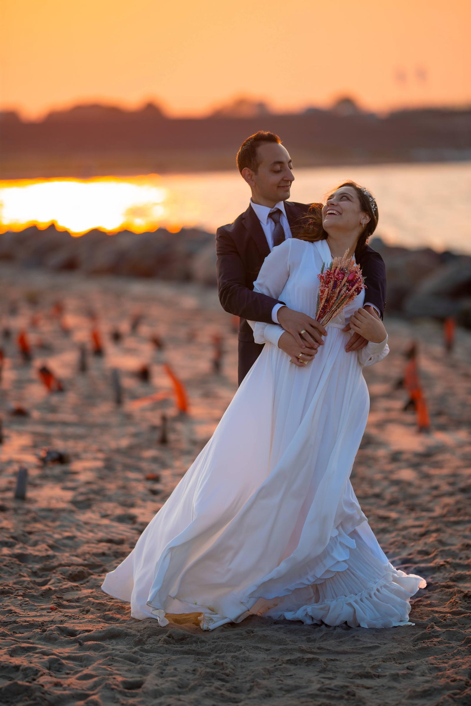 A couple in wedding attire embracing on a sandy beach at sunset. The woman is smiling while holding a bouquet, and the man is looking at her. The sky is orange and pink with the sun setting over water in the background.