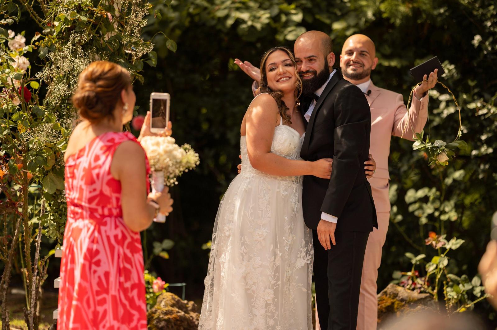 A bride and groom embrace during their wedding ceremony outdoors, with friends taking photos, surrounded by greenery and flowers.