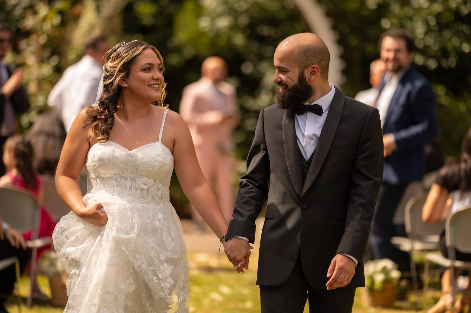 A bride and groom holding hands at their wedding ceremony outdoors, smiling at each other. In the background, guests are seated and standing among trees.