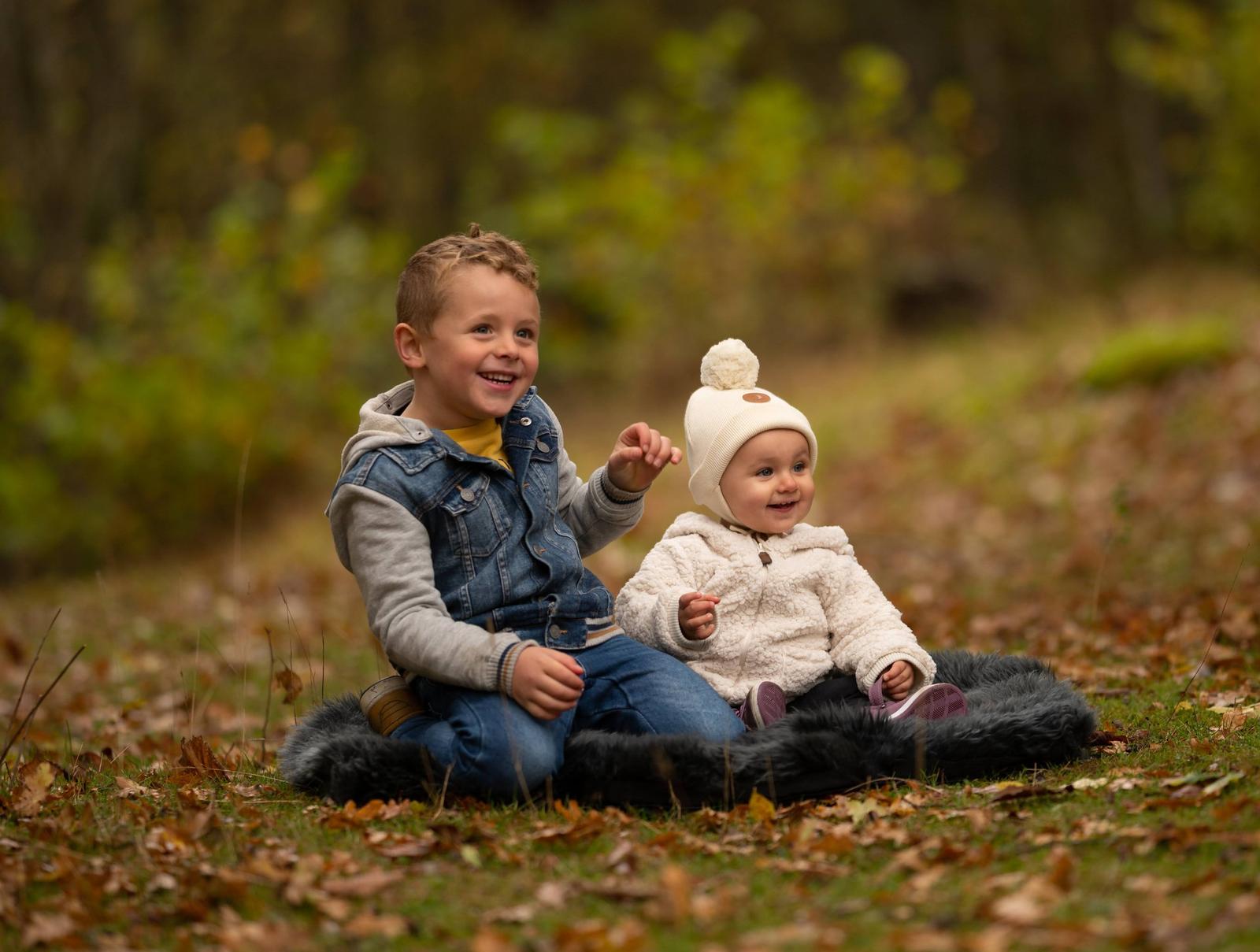 Two children, an older boy and a younger girl, sitting on a black fur blanket in an autumn forest, smiling and enjoying the outdoor scenery.