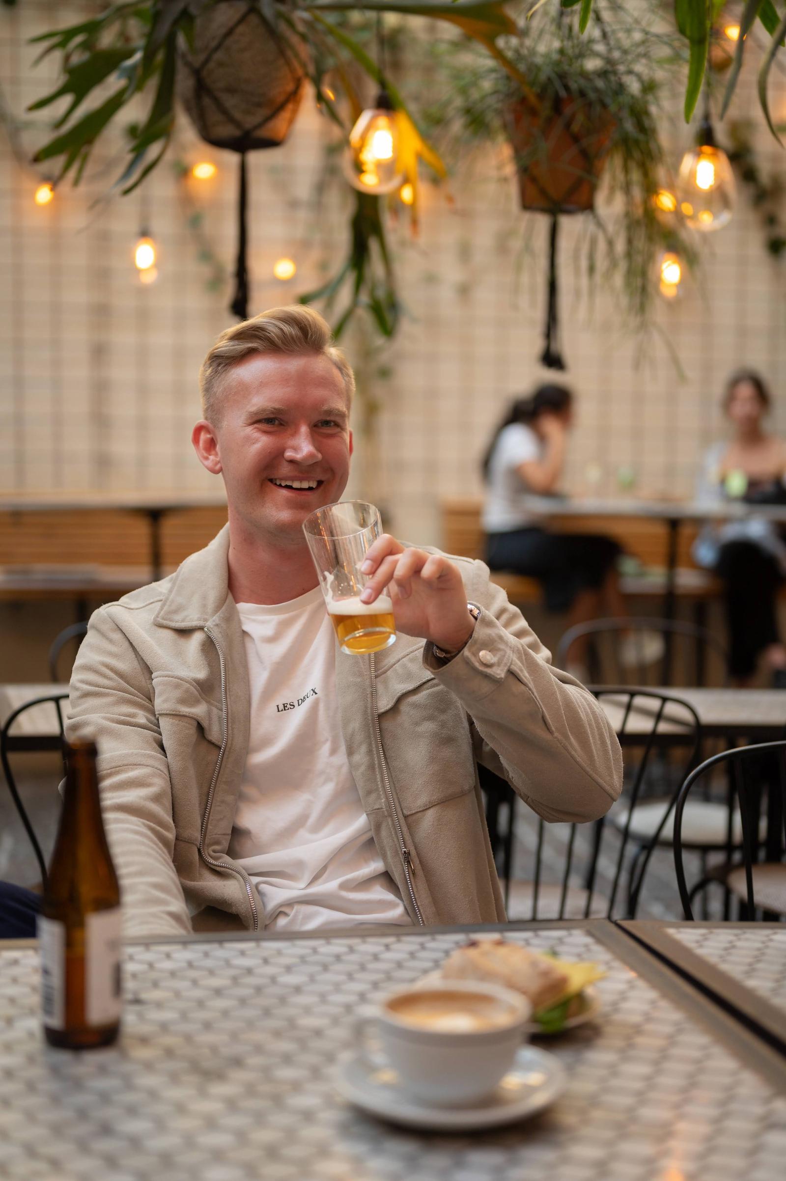 A young man with blond hair smiling and holding a glass of beer in a café or bar setting. In the foreground, there is a cup of coffee on a saucer and a beer bottle on the table. The background features warm lighting, hanging plants, and people sittin