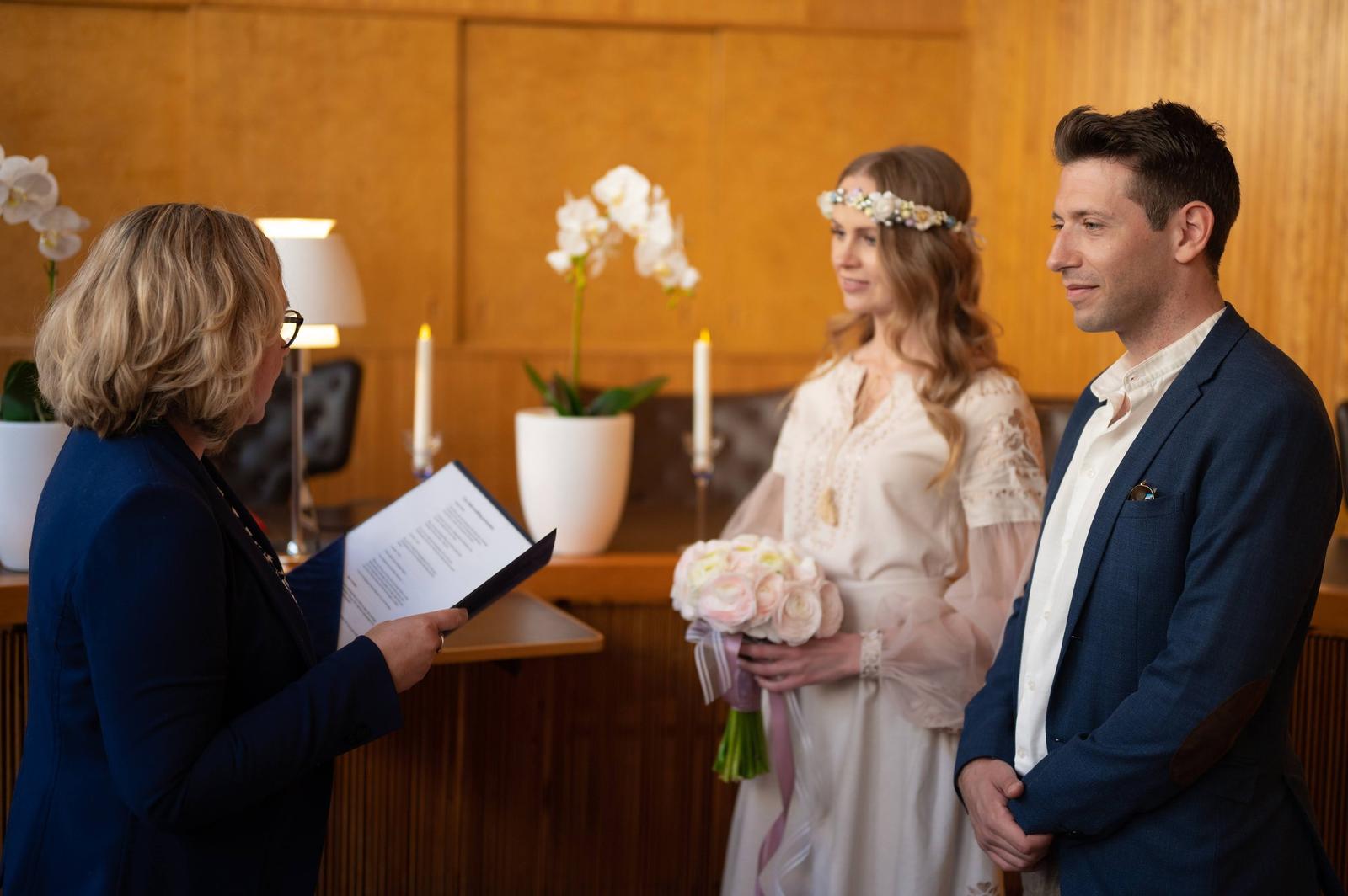 A wedding ceremony with a bride and groom standing before a woman reading from a document, in a warmly lit room decorated with white orchids and candles.