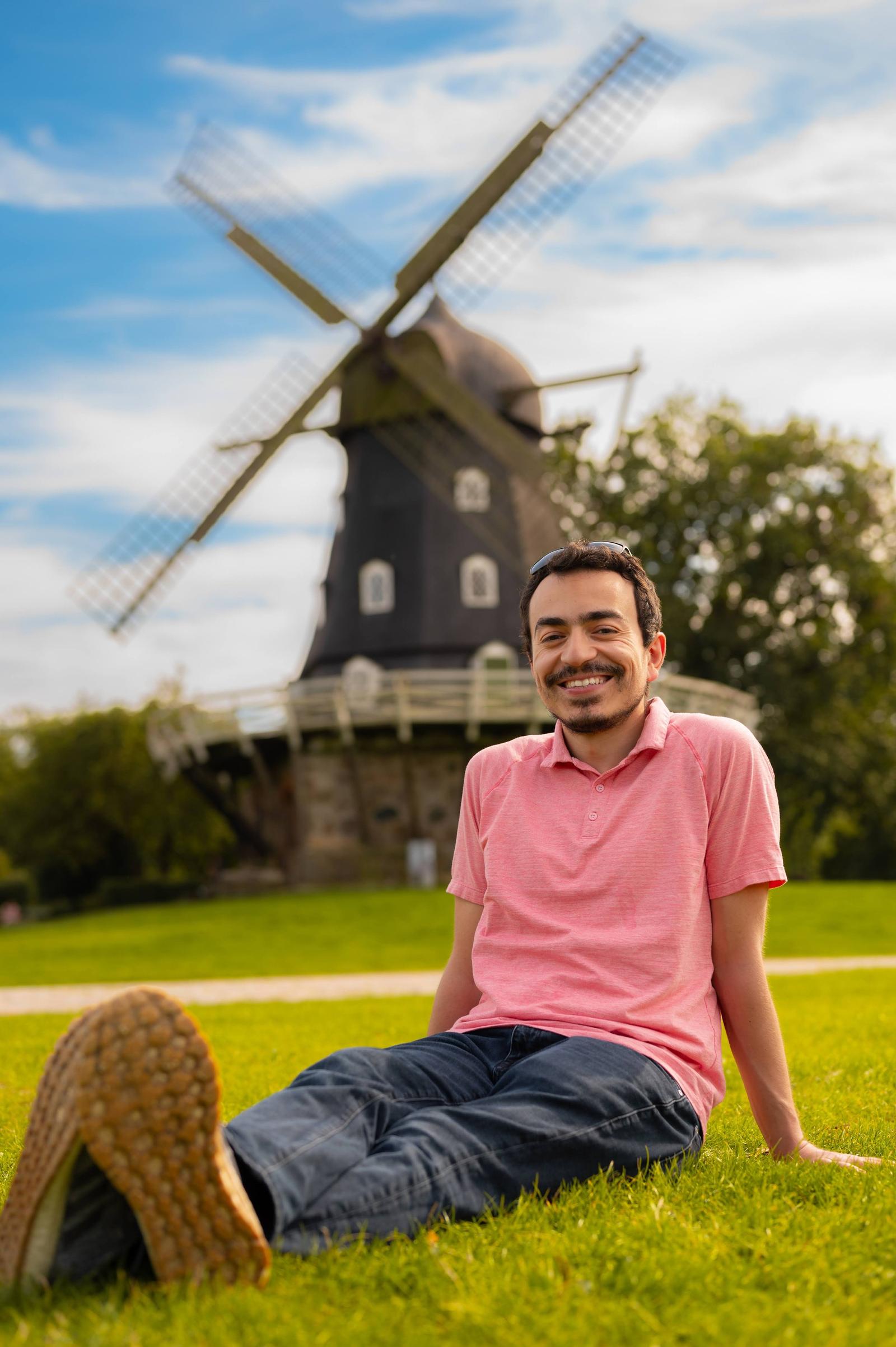 A young man sitting on the grass in front of a traditional windmill, smiling at the camera on a sunny day with blue sky and some clouds.