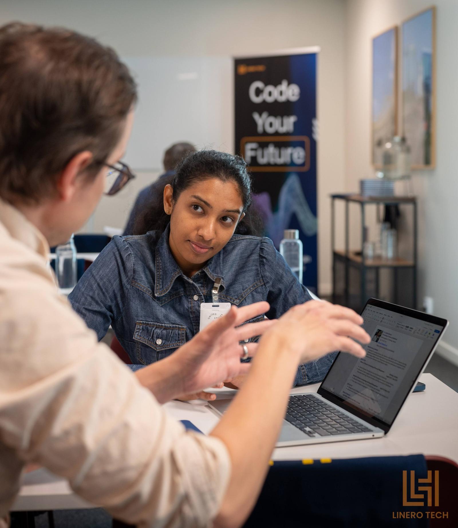 Two people in a meeting talking while looking at a laptop, in a room with a sign that says "Code Your Future".