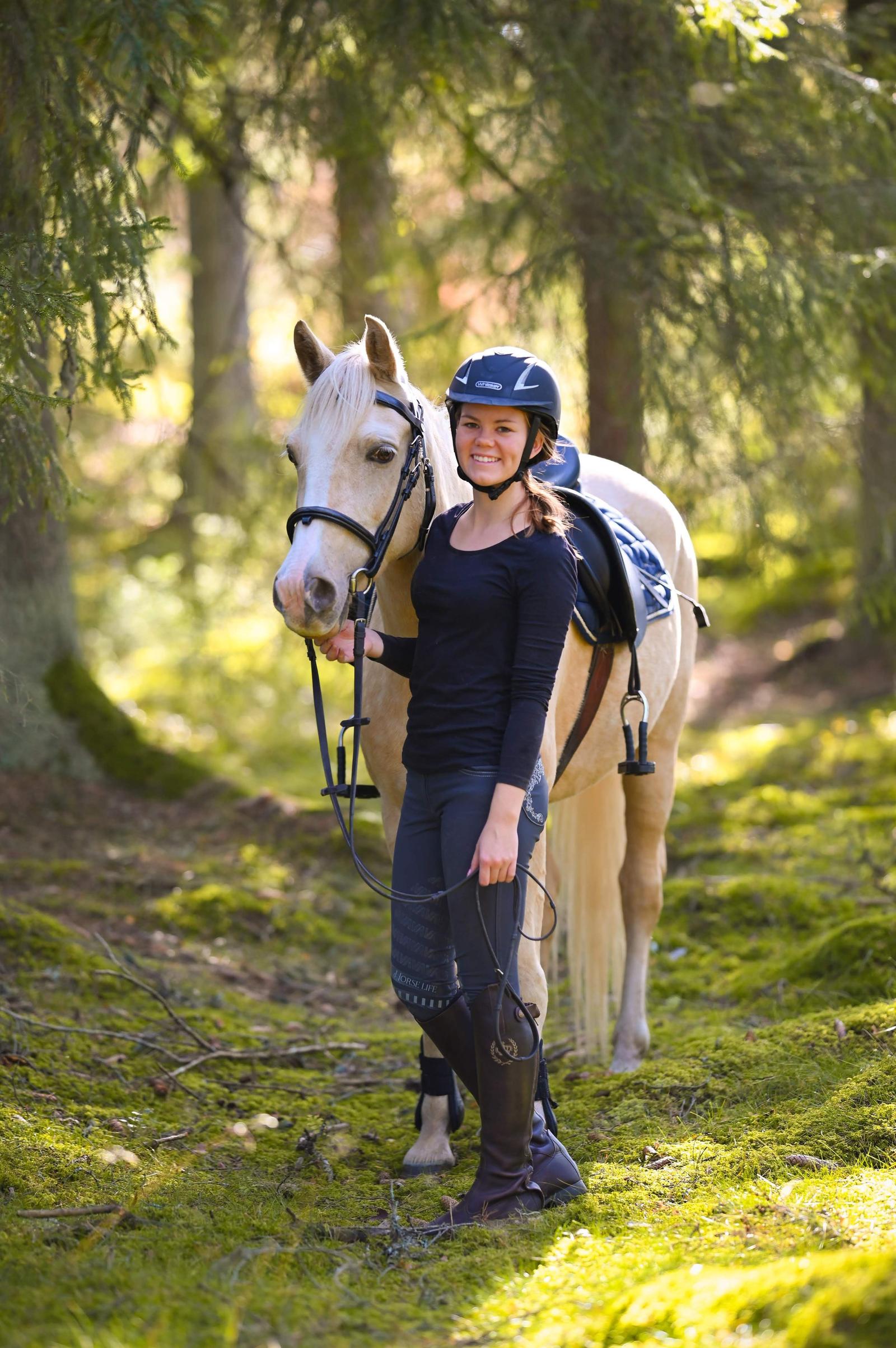 A young woman in riding gear stands with a white horse in a forest, smiling at the camera.