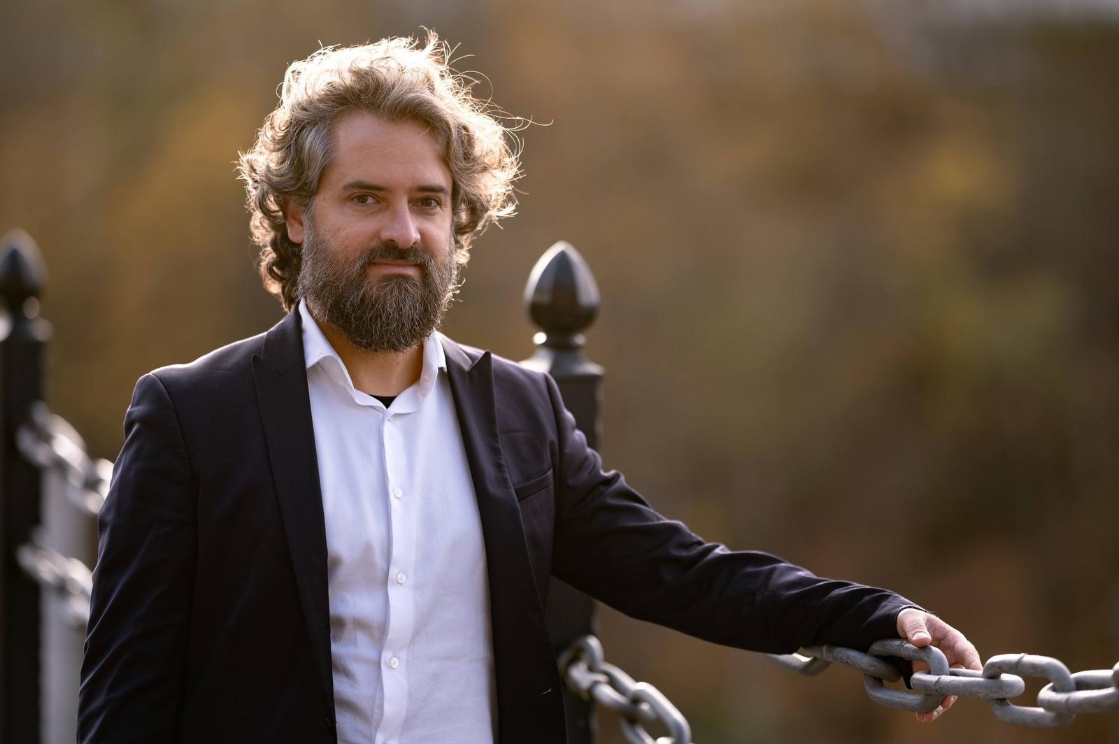 A man with gray curly hair and a beard, dressed in a white shirt and black blazer, standing outdoors on a bridge with his hand on a chain railing. The background is blurred with warm autumn colors.