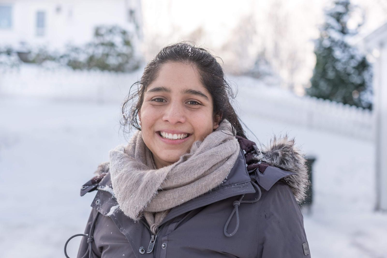 A young woman smiling outdoors in a snowy landscape, wearing a dark winter jacket, a beige scarf, and a fur-lined hood.