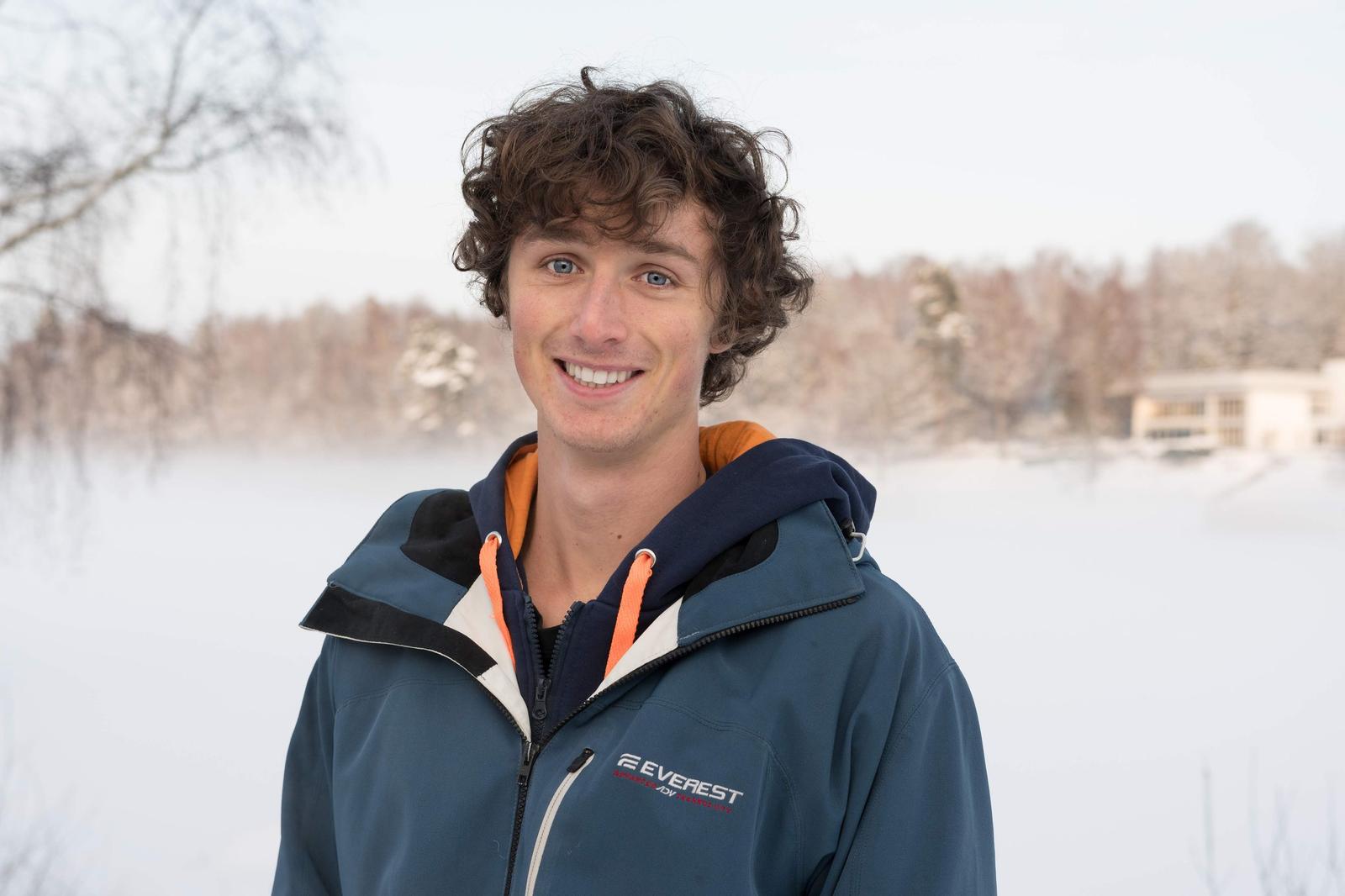 A young man with curly brown hair and blue eyes smiling outdoors in winter, wearing a blue and orange winter jacket and hoodie, with snow-covered trees and a snowy landscape in the background.