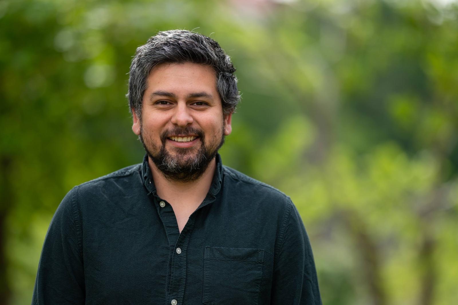 A man with dark hair and a beard smiling outdoors with a green, blurred background.