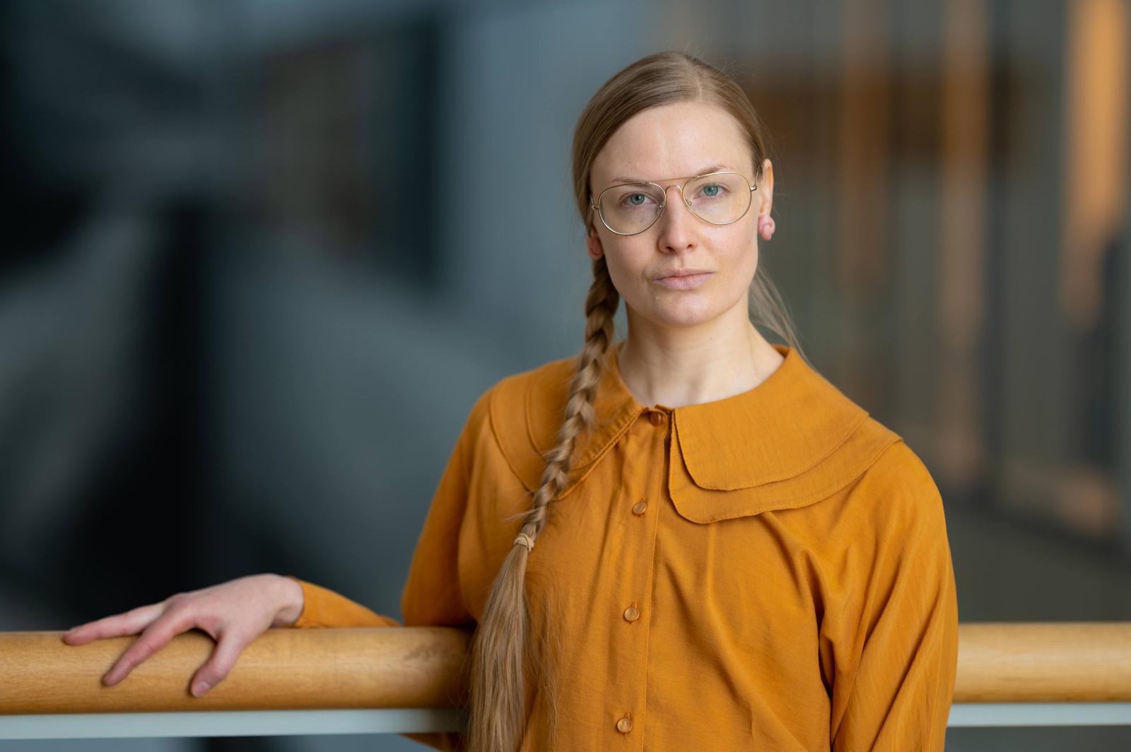 A woman with long braided blonde hair, wearing glasses and a mustard yellow blouse, standing indoors next to a wooden railing, looking at the camera.