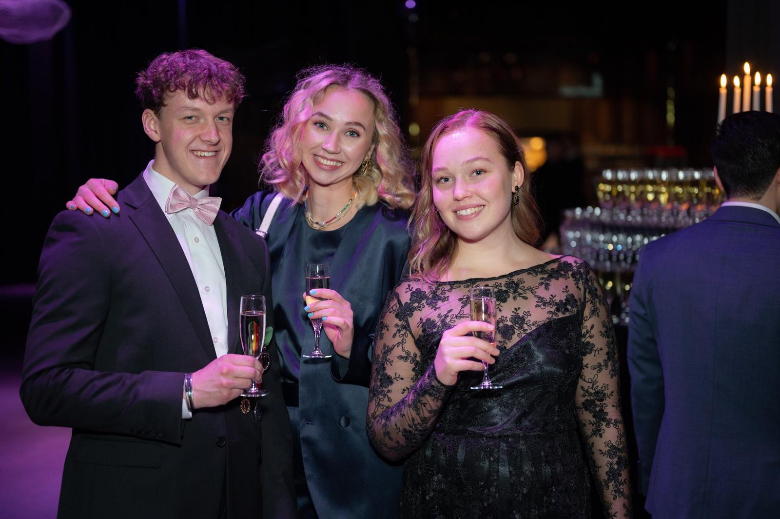 Three young adults dressed in formal attire at a celebration, holding glasses of champagne and smiling for the camera, in an indoor setting with dim lighting and candles.
