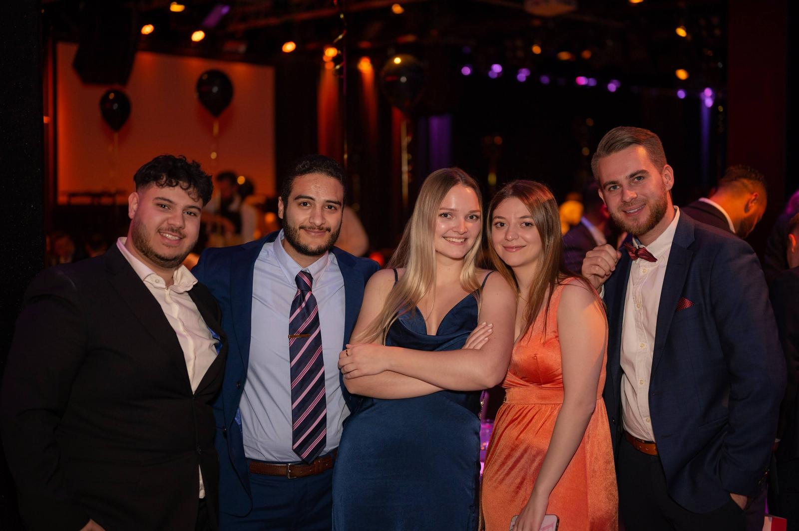 A group of five young adults dressed formally at an indoor event with colorful lighting and decorations.