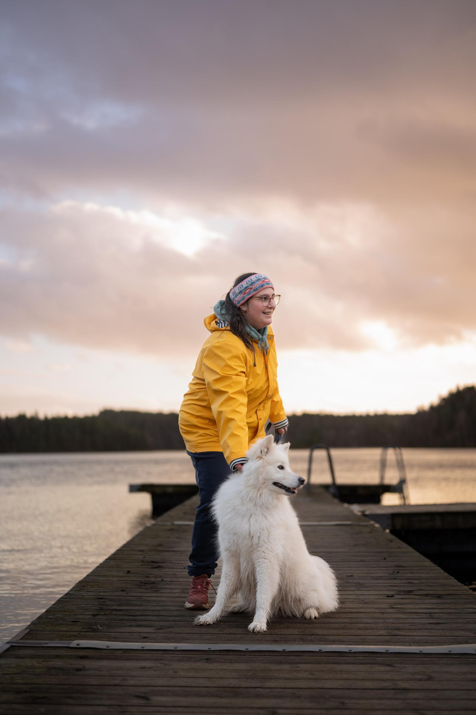 A woman in a yellow jacket and glasses standing on a dock next to a white fluffy dog by a lake during sunset.