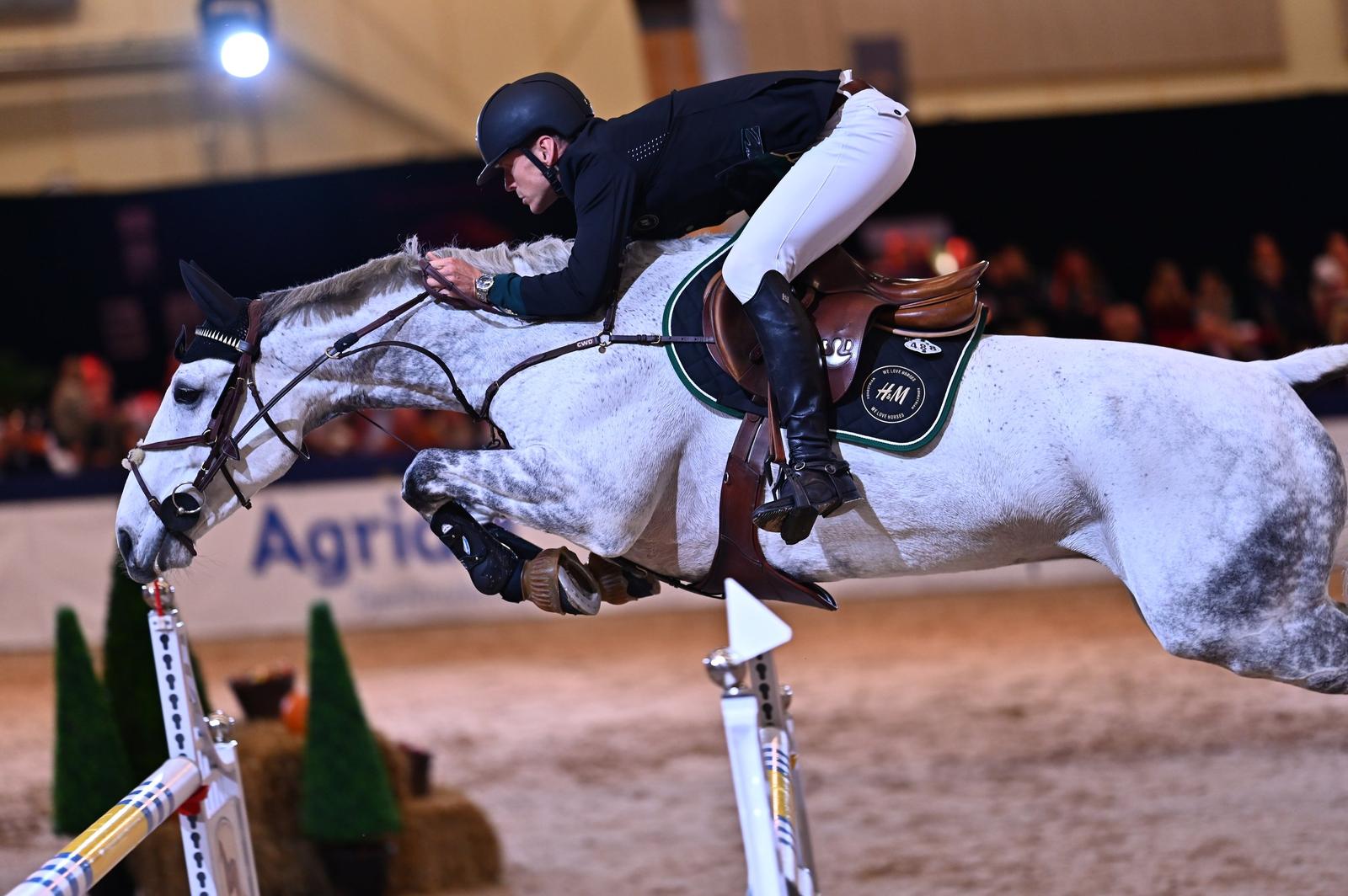 A male equestrian athlete wearing a black helmet, black jacket, and white riding pants, riding a grey horse as they clear a jump in an indoor arena during a show jumping competition.