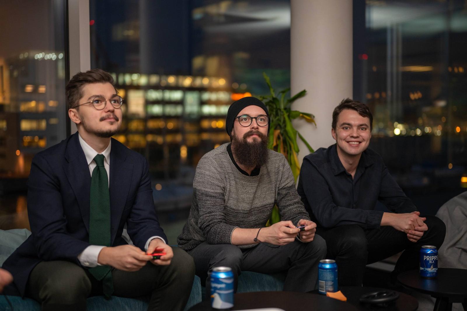 Three men sitting on a couch in a modern office or lounge area, playing video games at night with city lights visible through large windows behind them.