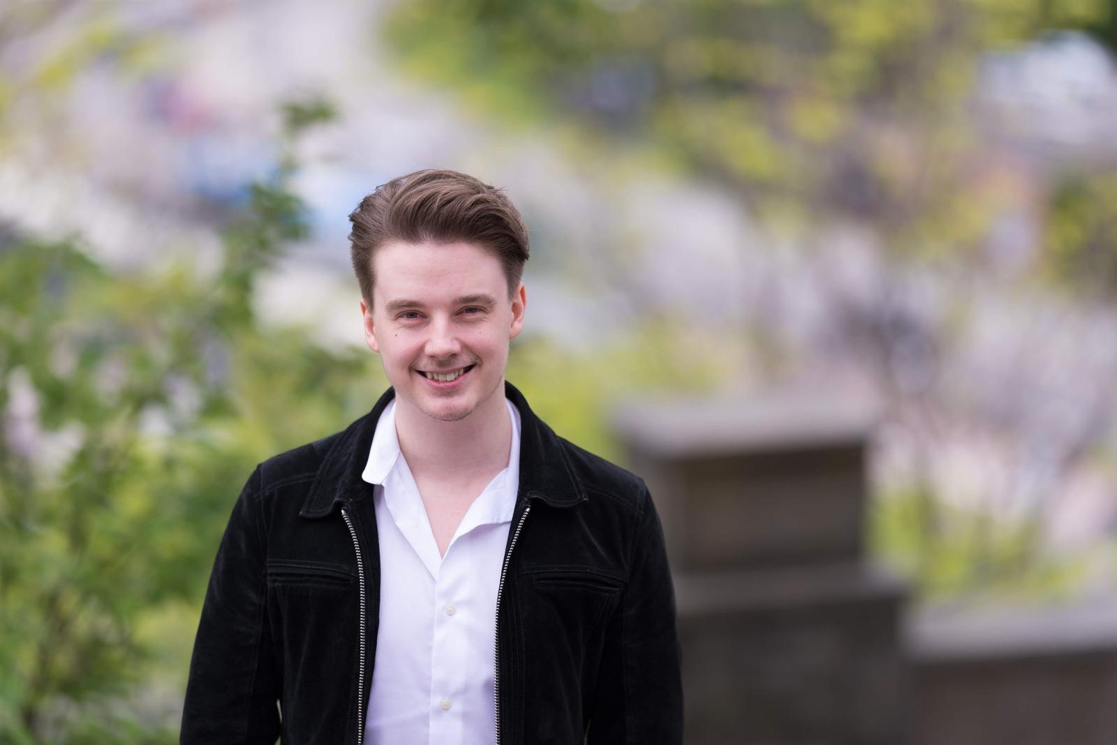 Young man with brown hair smiling outdoors, wearing a white shirt and black jacket, with blurred trees and steps in the background.