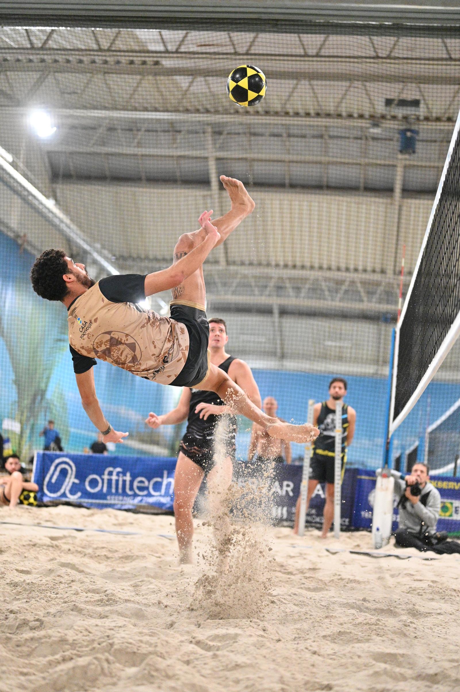 A beach volleyball player dives mid-air to hit the ball, with spectators and other players in the background on a sand court.