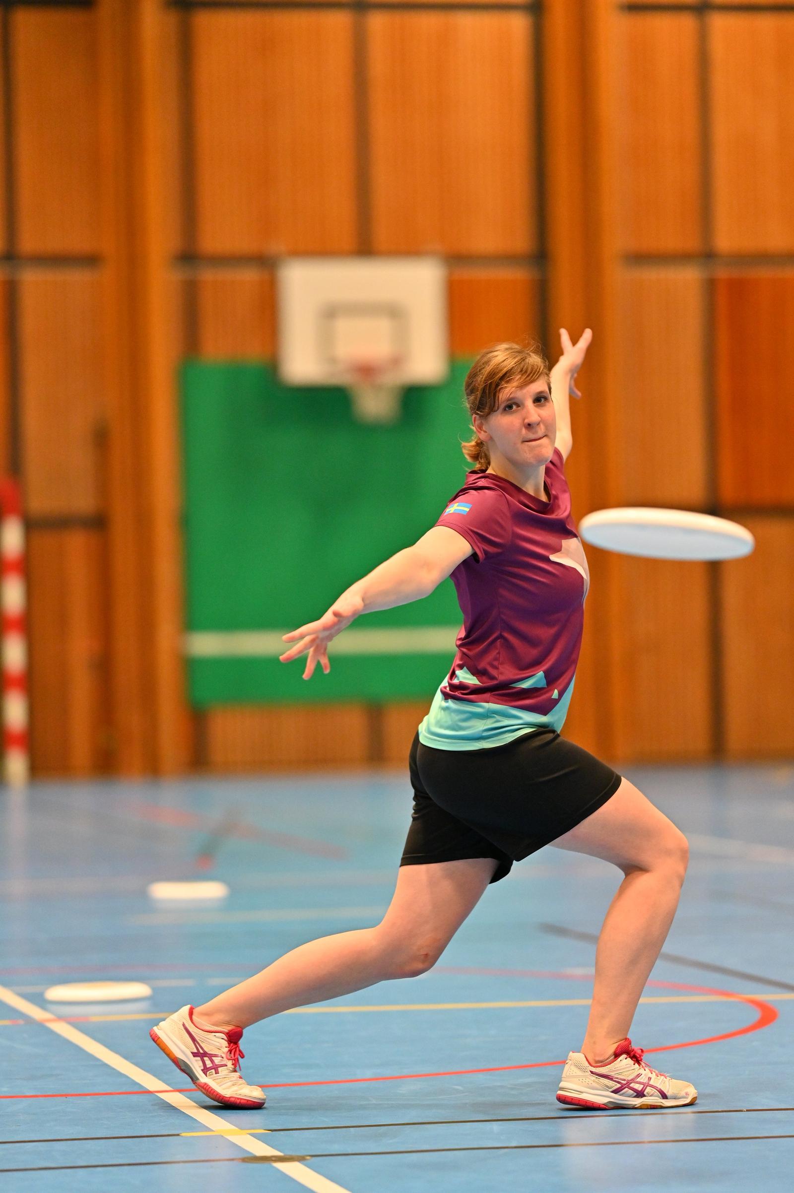 A woman playing ultimate frisbee in an indoor gym, wearing a maroon and teal sports shirt, black shorts, and white sneakers, with a frisbee in front of her.