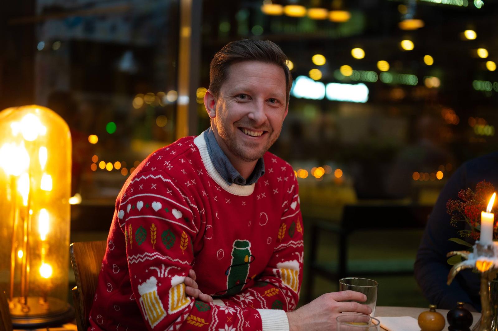 Man smiling at a festive dinner table during an evening celebration, wearing a Christmas sweater with holiday designs, illuminated by candlelight and warm decorative lighting in the background.