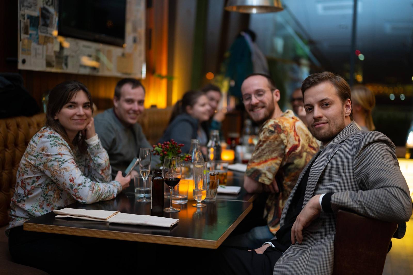 People sitting at a restaurant table with drinks and a floral centerpiece, smiling at the camera.