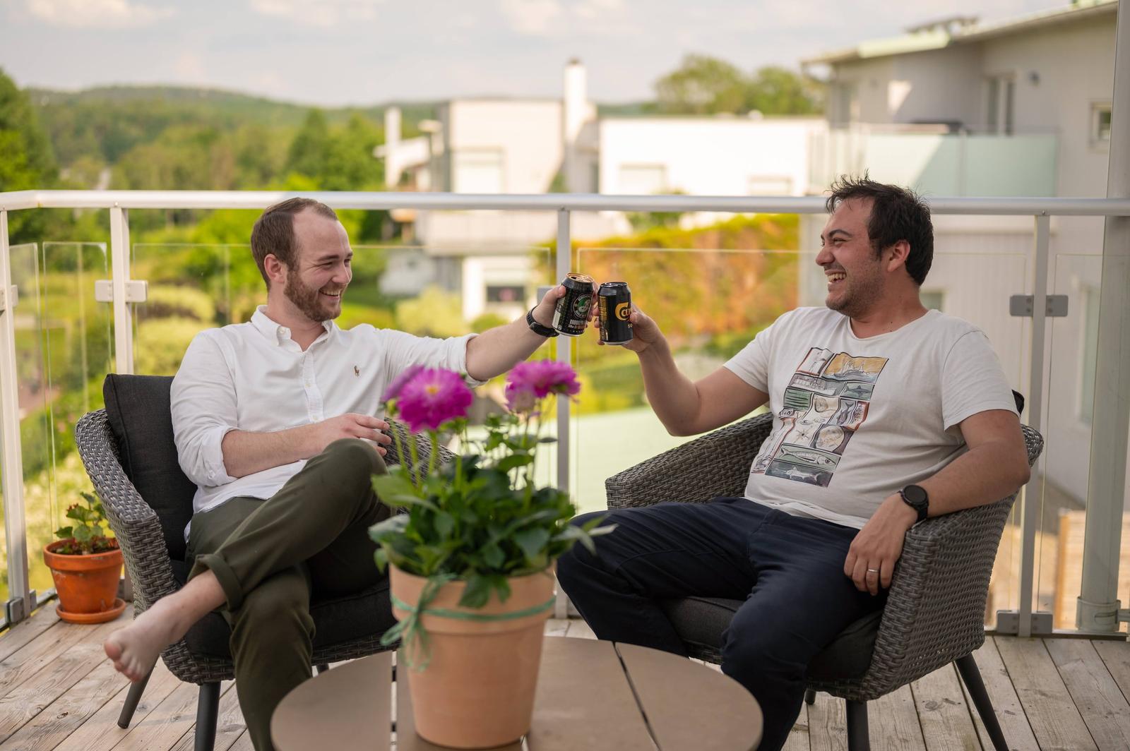 Two men sitting on a balcony, smiling and clinking cans of soda in a toast. One man has a white shirt and brown pants, the other has a white t-shirt with a graphic design and dark pants. Potted plants and a scenic background with green hills are visi