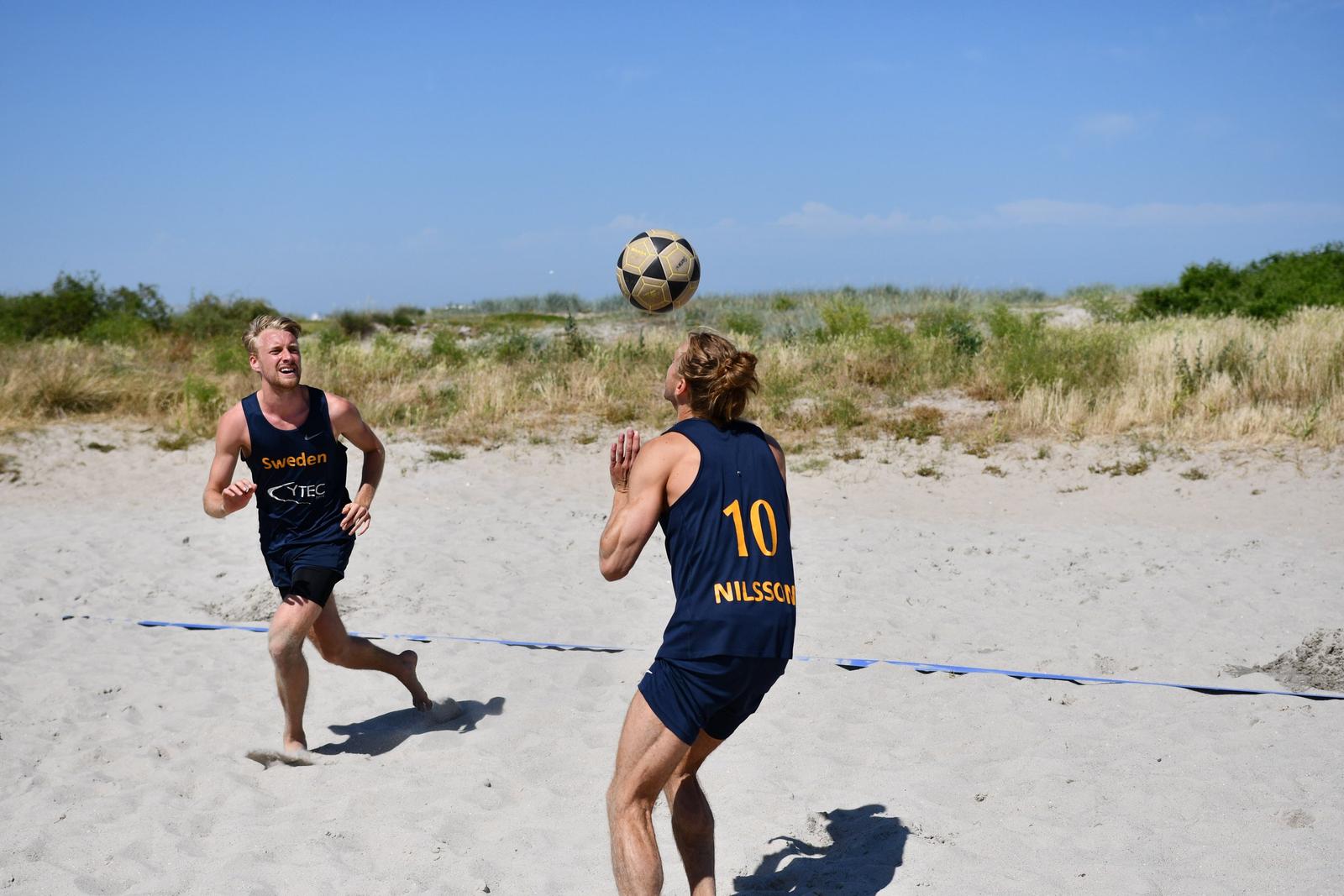 Two young men playing beach soccer on sand with grassy dunes in the background, one about to catch a soccer ball in mid-air.