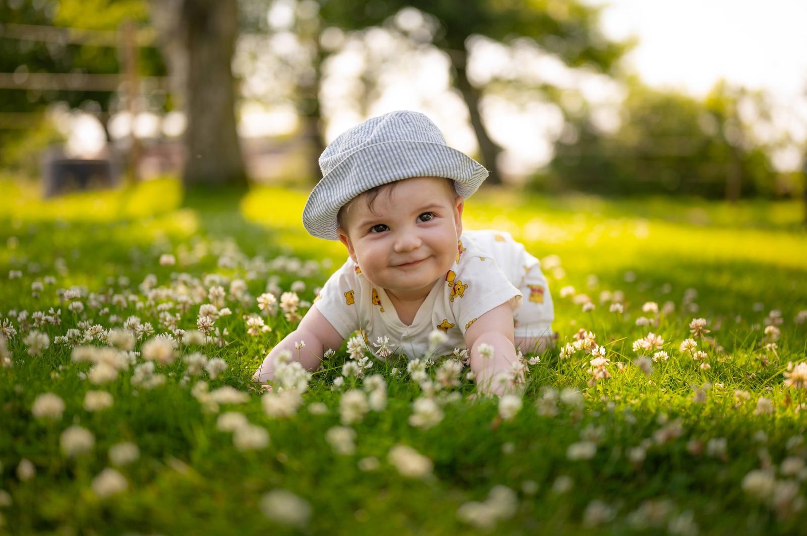 A baby crawling on grass in a park, wearing a striped hat and a white shirt with yellow bear prints, surrounded by small white flowers with trees and sunlight in the background.