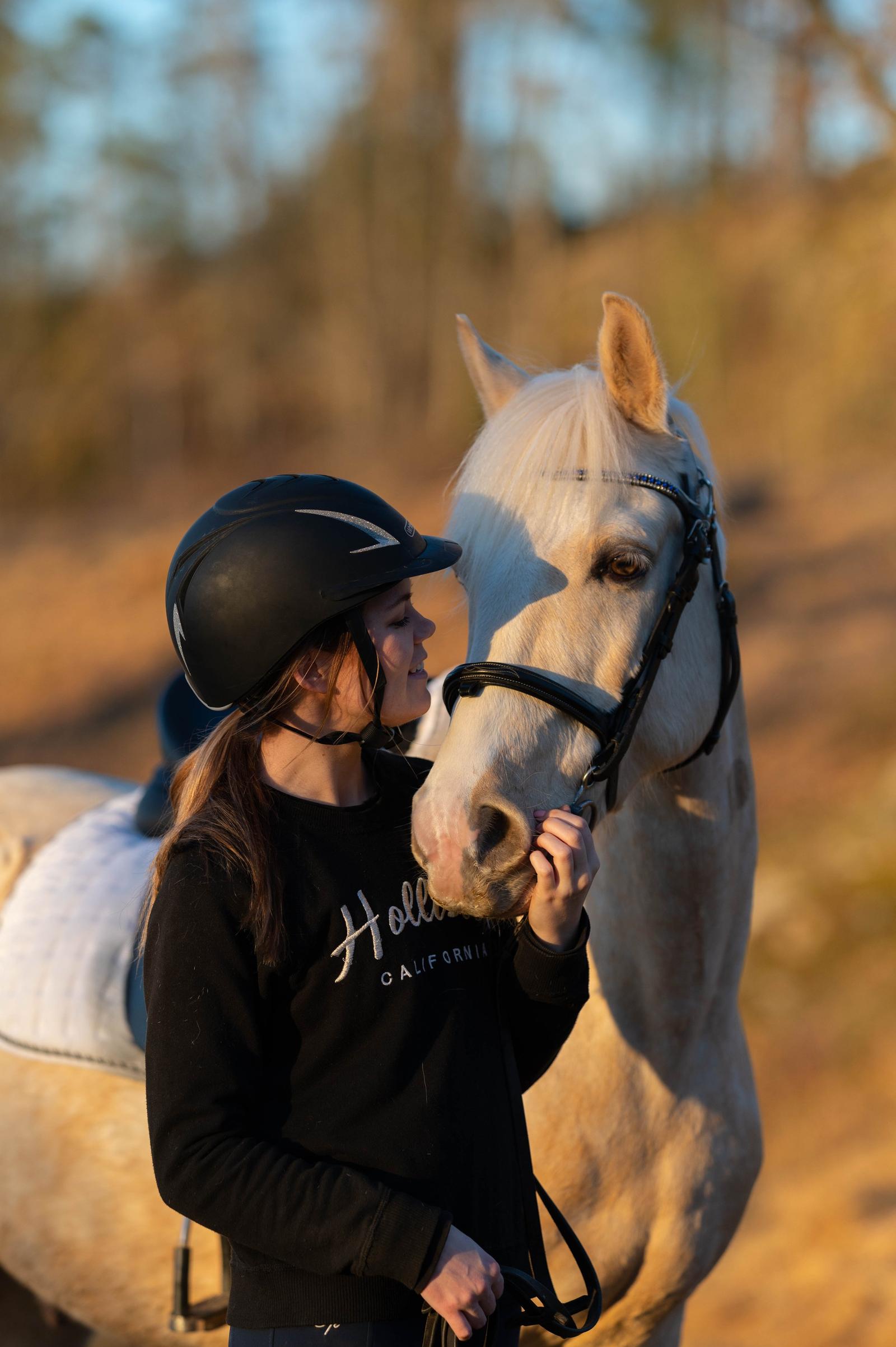 A young woman wearing a black helmet and sweatshirt petting a white horse outdoors during sunset.