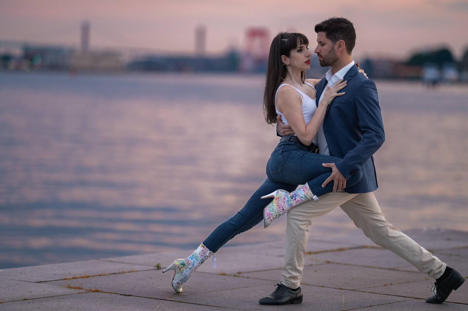 A couple dancing by the water at sunset, the woman wearing colorful high-heeled boots and the man dressed in a blue blazer and beige pants.