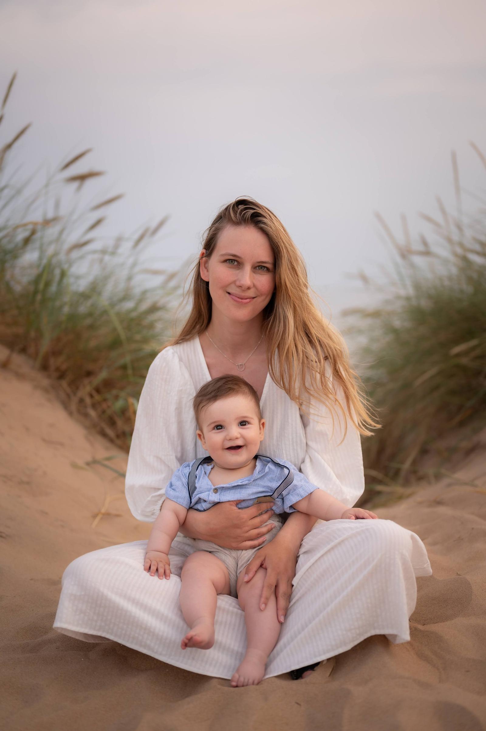 A woman and a baby sitting on a sandy beach with tall grass in the background, both smiling at the camera.