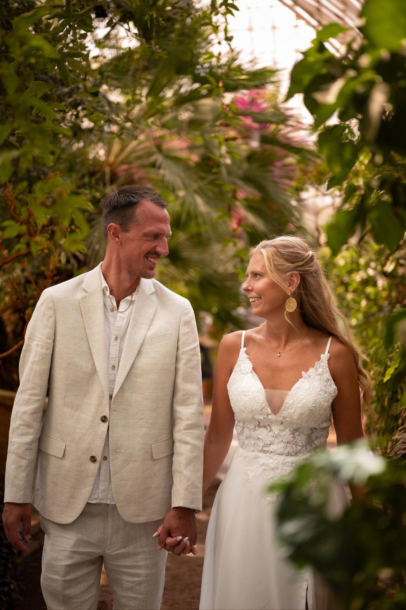 A bride and groom holding hands and smiling at each other in a lush, green environment.