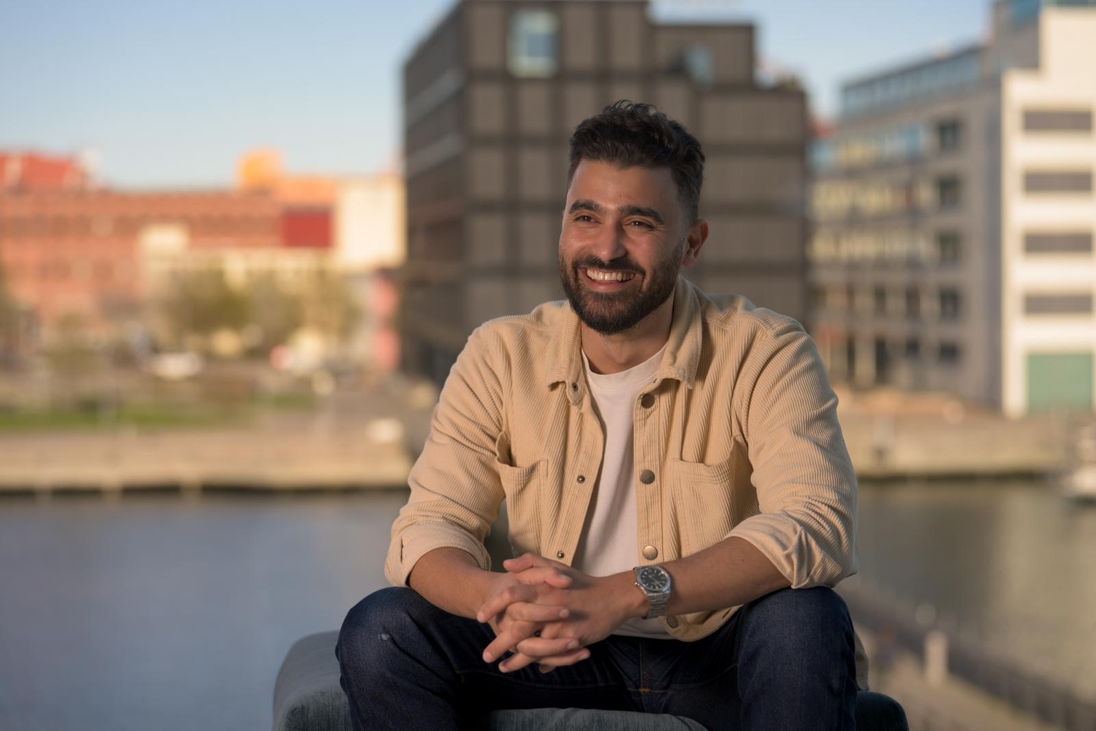 Portrait of a smiling bearded man in a beige corduroy shirt sitting by the water in the evening sun, with city buildings in the background