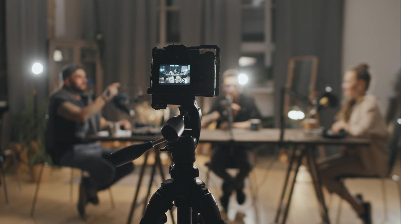 A camera on a tripod filming a group of people sitting around a table during a meeting or interview indoors, with a blurred background.