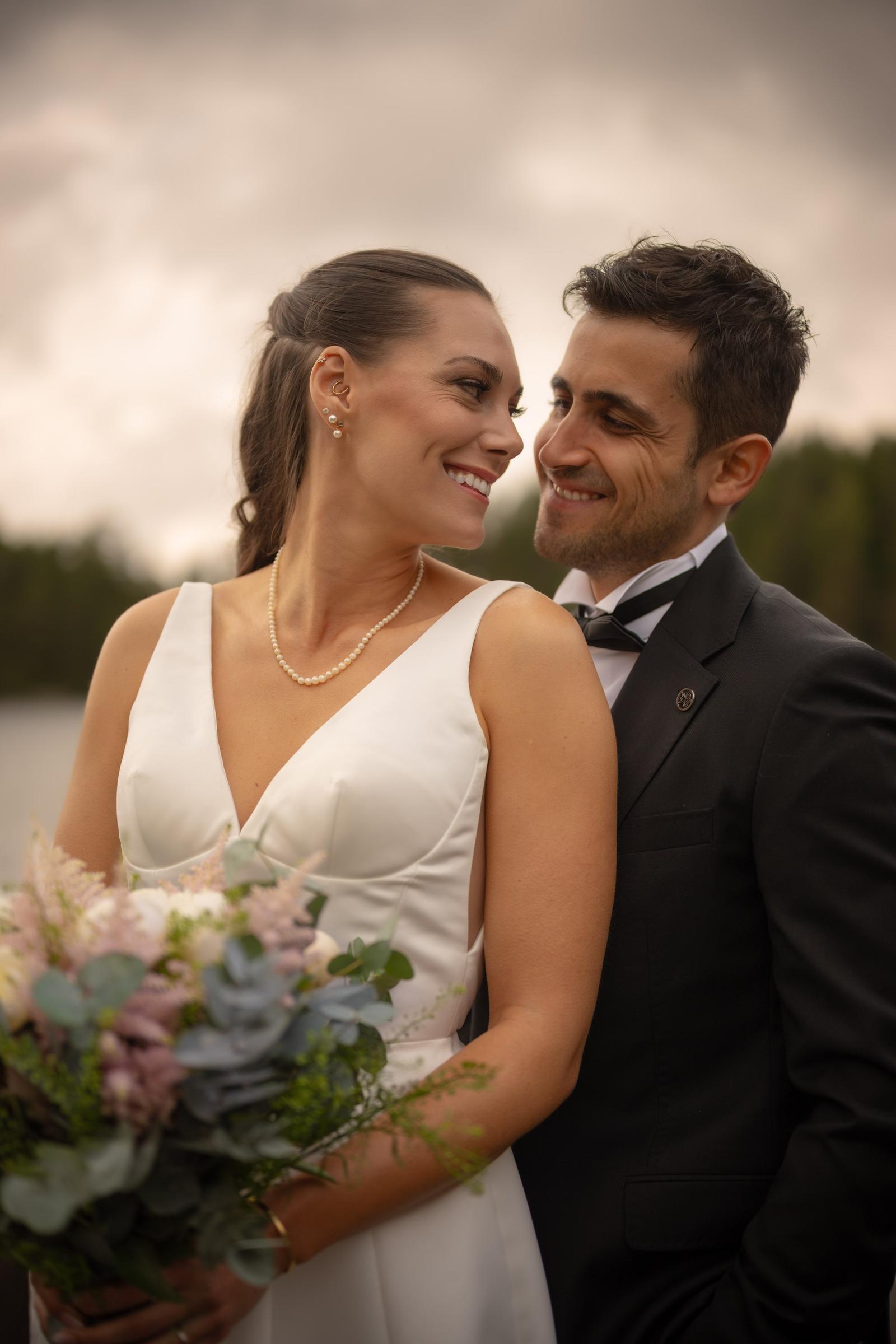 Newlyweds smiling at each other outdoors with a beautiful natural landscape in the background.