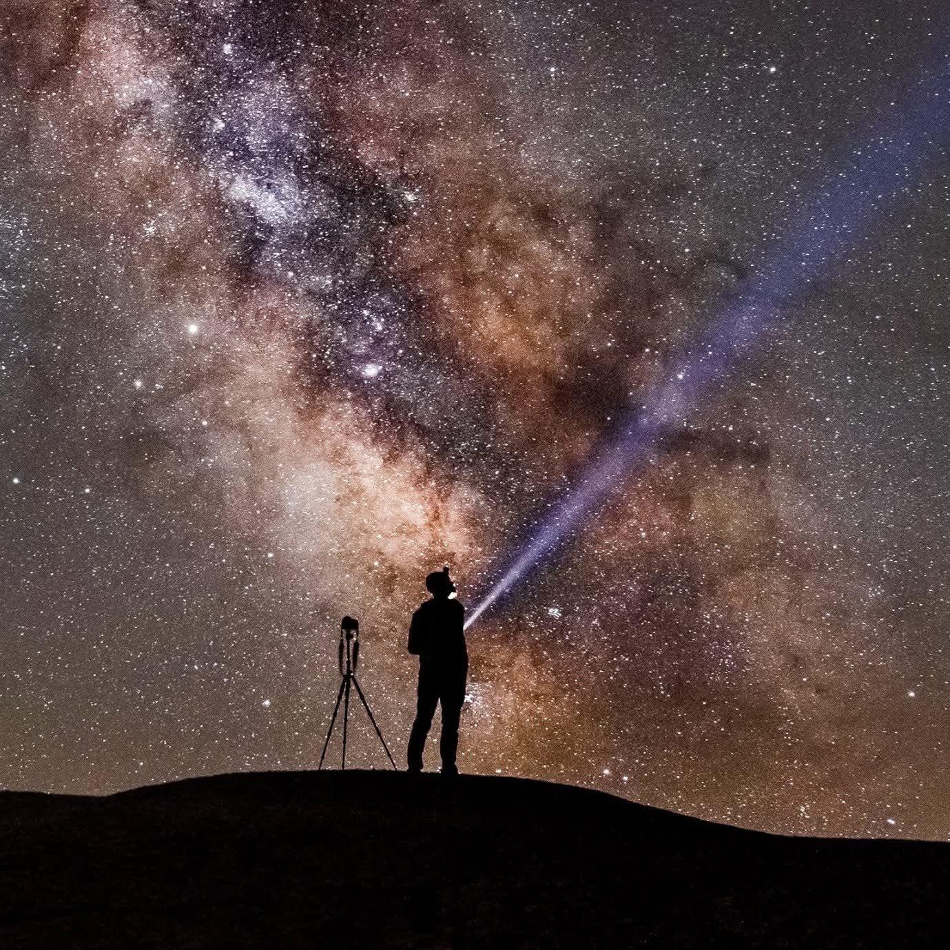 A person standing on a mountain top, pointing a torch at the stars with the Milky Way across the sky and a tripod next to them.
